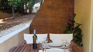 Mesa de madera para dos personas, con una botella de vino, copas y platos, en un patio con vistas a la vegetación y una terraza de madera.