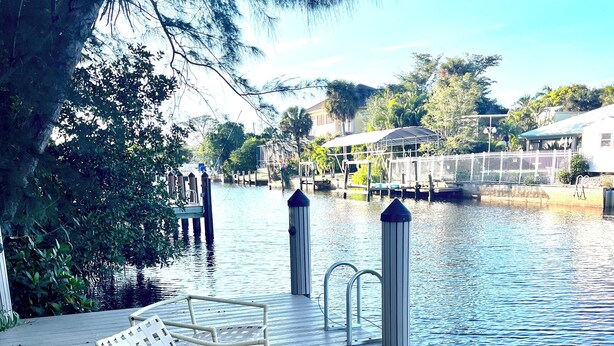 A wooden deck with two wicker chairs, a metal table, and a dock extending into a calm body of water. - Relaxing house with spacious dock tucked on Gordon River. Live the Florida dream (Naples)