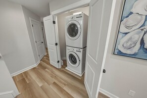 A laundry room with two stacked washers and dryers, wooden flooring, and a painting on the wall. - HOT TUB! Four bedroom home in the 4X4, Corolla, NC. A truly unique getaway! (Corolla)