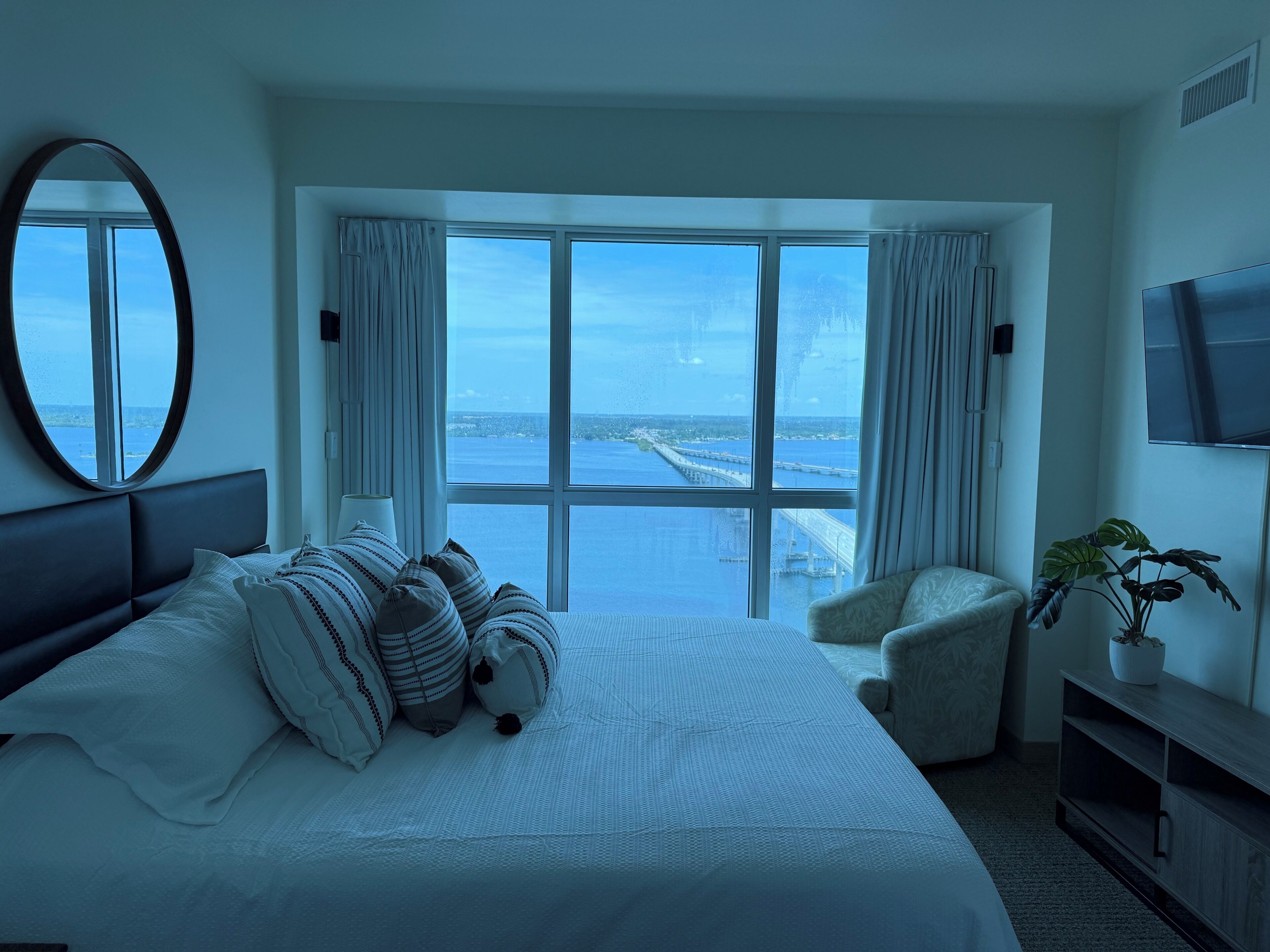 A hotel room with a large bed, a chair, a TV, and a view of the sea and a bridge.