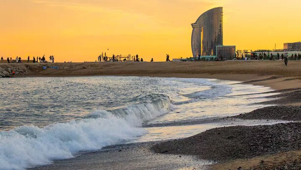 Ein Strand mit Wellen bei Sonnenuntergang, ein gebogenes Gebäude und Personen in der Ferne.