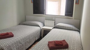 Ein Zimmer mit zwei Betten, beide mit gemusterten Bettbezügen und einem roten Handtuch. Ein Fenster mit Fensterläden, ein Heizkörper und ein kleiner Tisch zwischen den Betten.