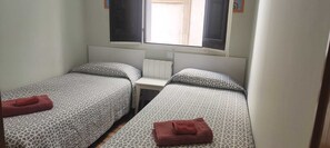 A room with two beds, each with patterned bedding and a red towel. There is a window with shutters, a radiator, and a small table between the beds. - Acomodis great location right in El Born old town 4 air conditioners 6 ppl max.! (Barcelona)