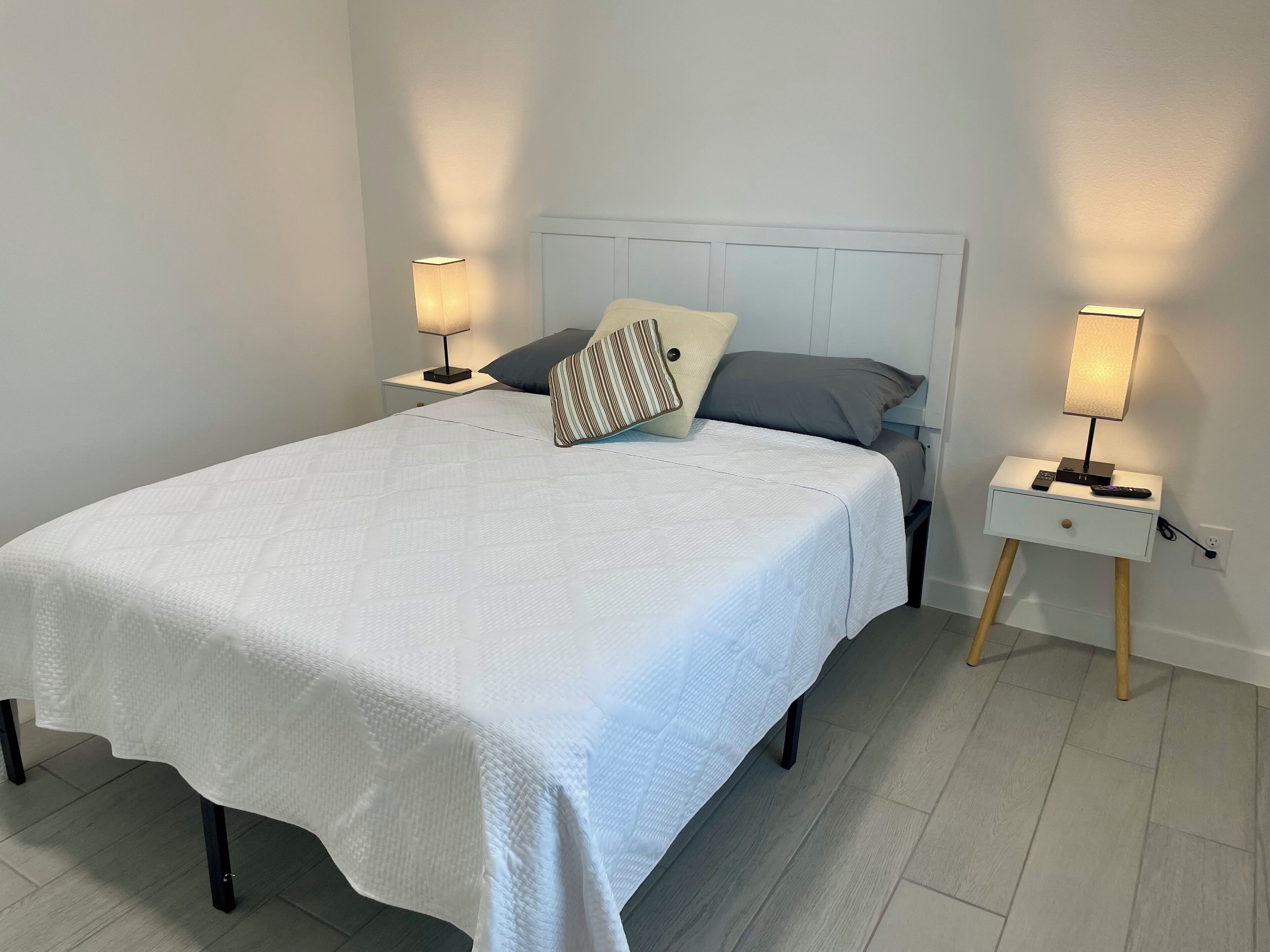 A neatly made bed with a white bedspread, two bedside tables with lamps, and a wooden nightstand with a drawer.