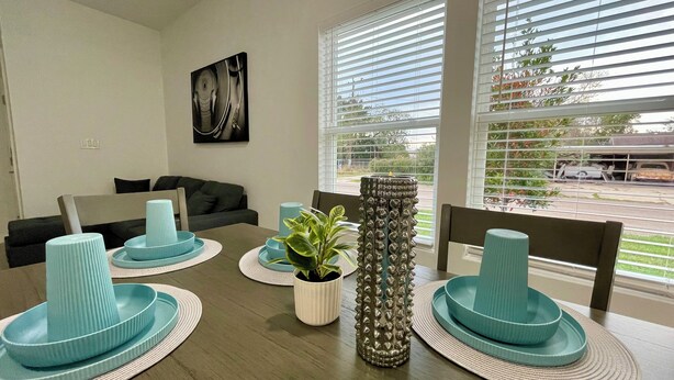 Comedor con una mesa de madera dispuesta con platos y cuencos azul verdoso, una planta en maceta y un florero decorativo.