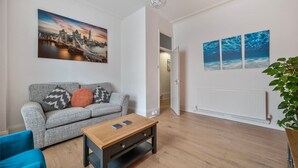 Un salon moderne avec un canapé gris, une table basse en bois et un fauteuil bleu. Un tableau représentant un paysage urbain est accroché au mur, et une plante se trouve dans un coin.