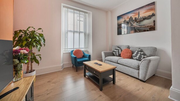 Un salon avec un canapé gris, un fauteuil bleu, une table basse en bois et un tableau représentant un paysage urbain.