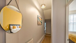 Un couloir avec un miroir rond, un radiateur, un tableau accroché au mur, et un lit avec une tête de lit jaune dans la pièce adjacente.