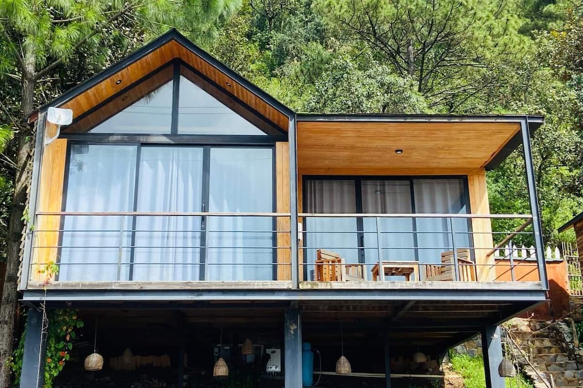 A modern two-story cabin with large windows and a balcony, surrounded by lush greenery.