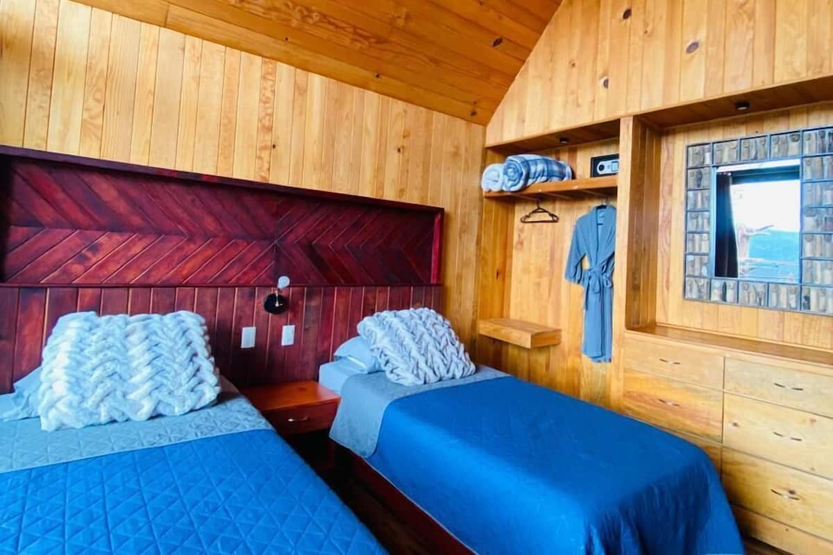 A wooden cabin room with two beds, a red herringboard headboard, a small window with a view, and a wooden dresser.
