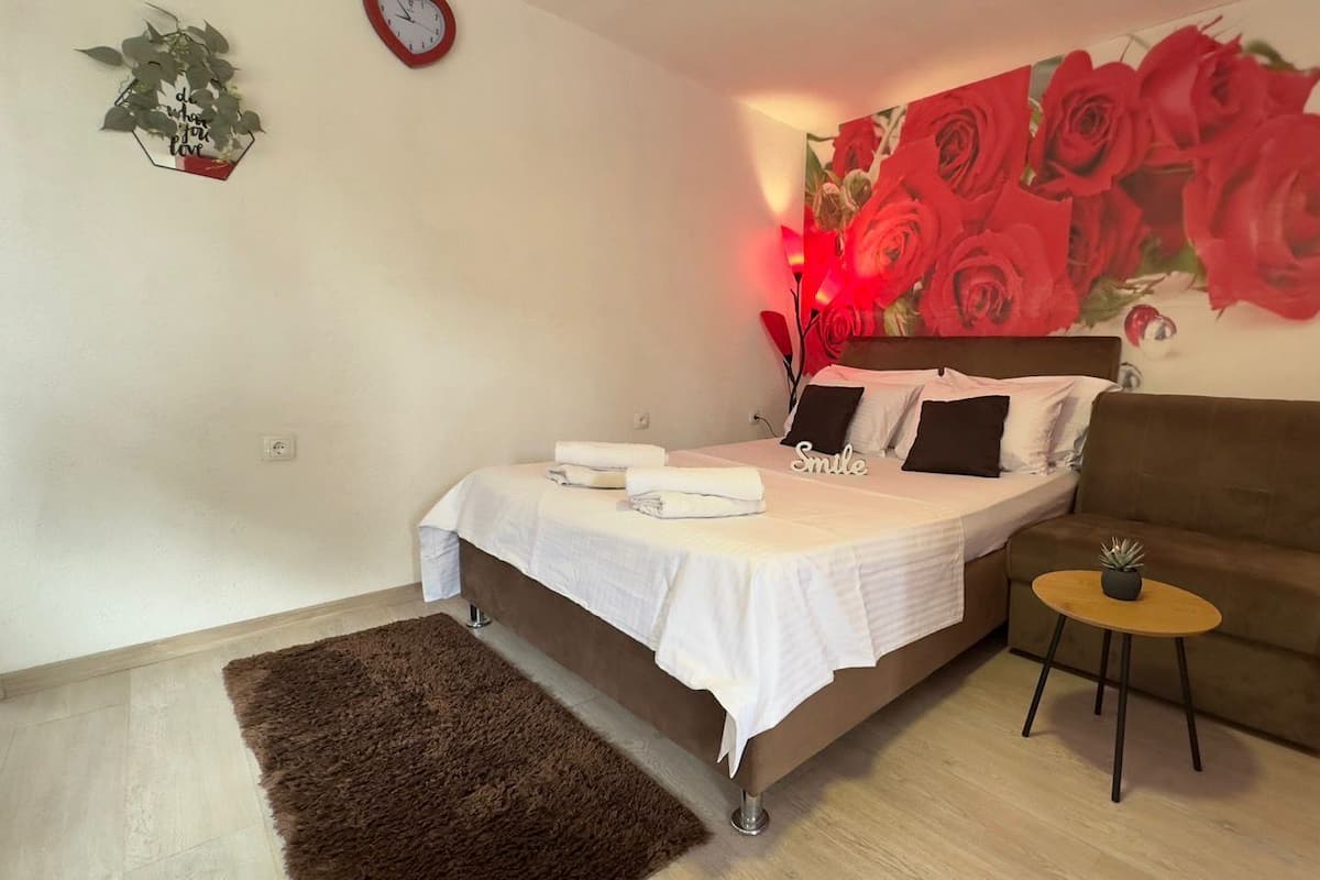 A bedroom with a bed, a brown armchair, a small round table, a wall clock, and a red rose painting.