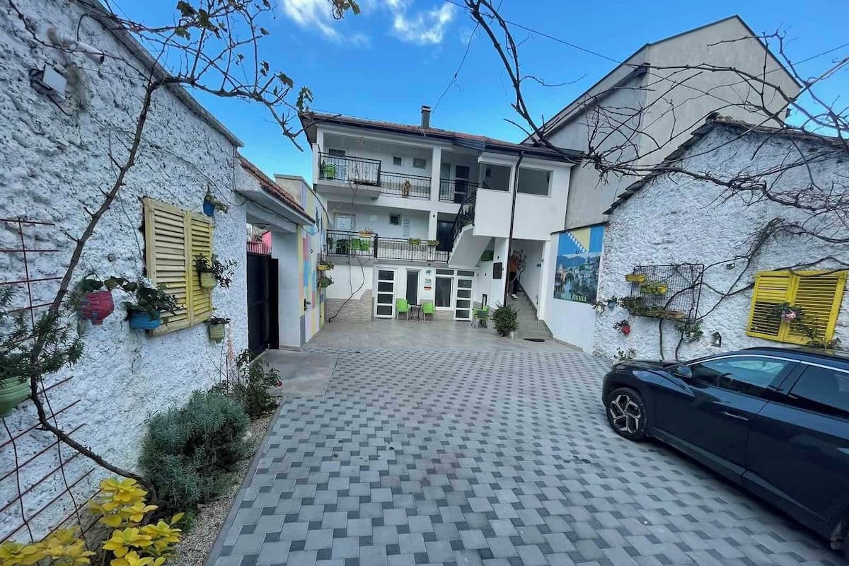 A courtyard with a tiled floor, a black car parked on the right, and a building with balconies and potted plants.