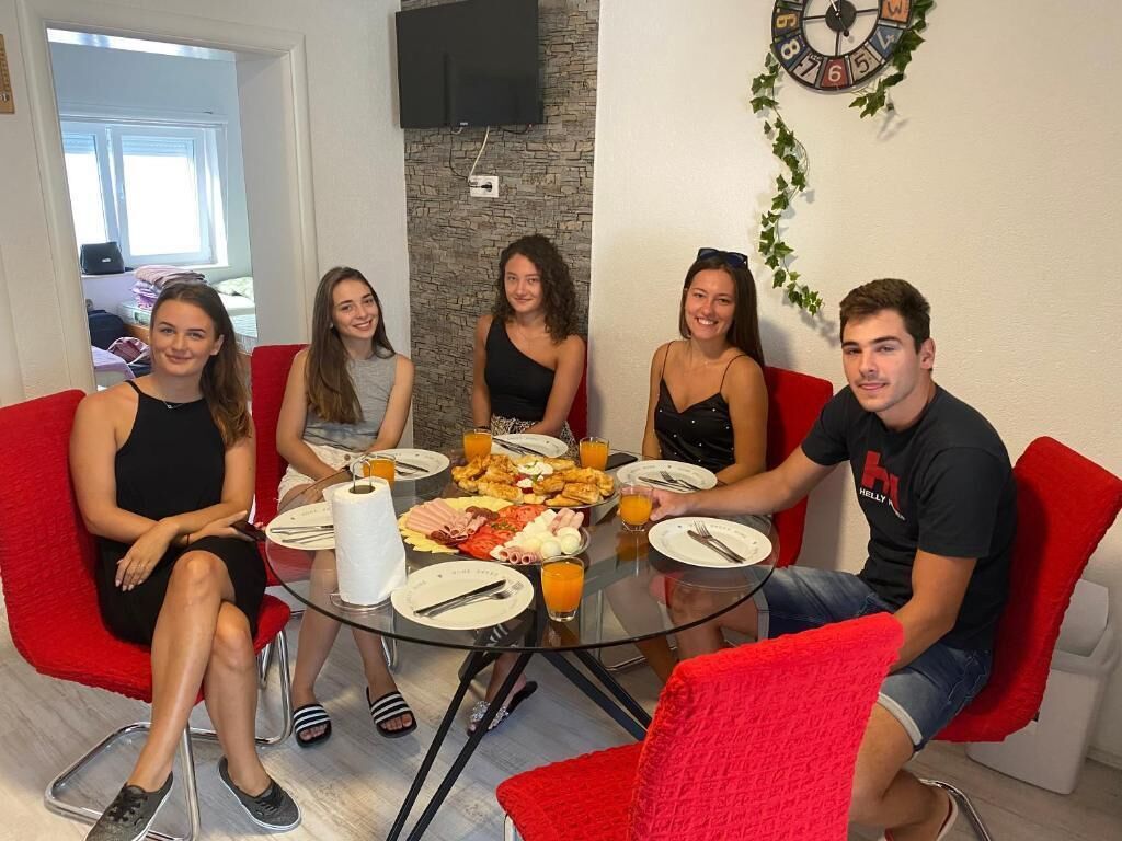 A group of people seated around a glass table with food and drinks, in a room with red chairs and a clock on the wall.