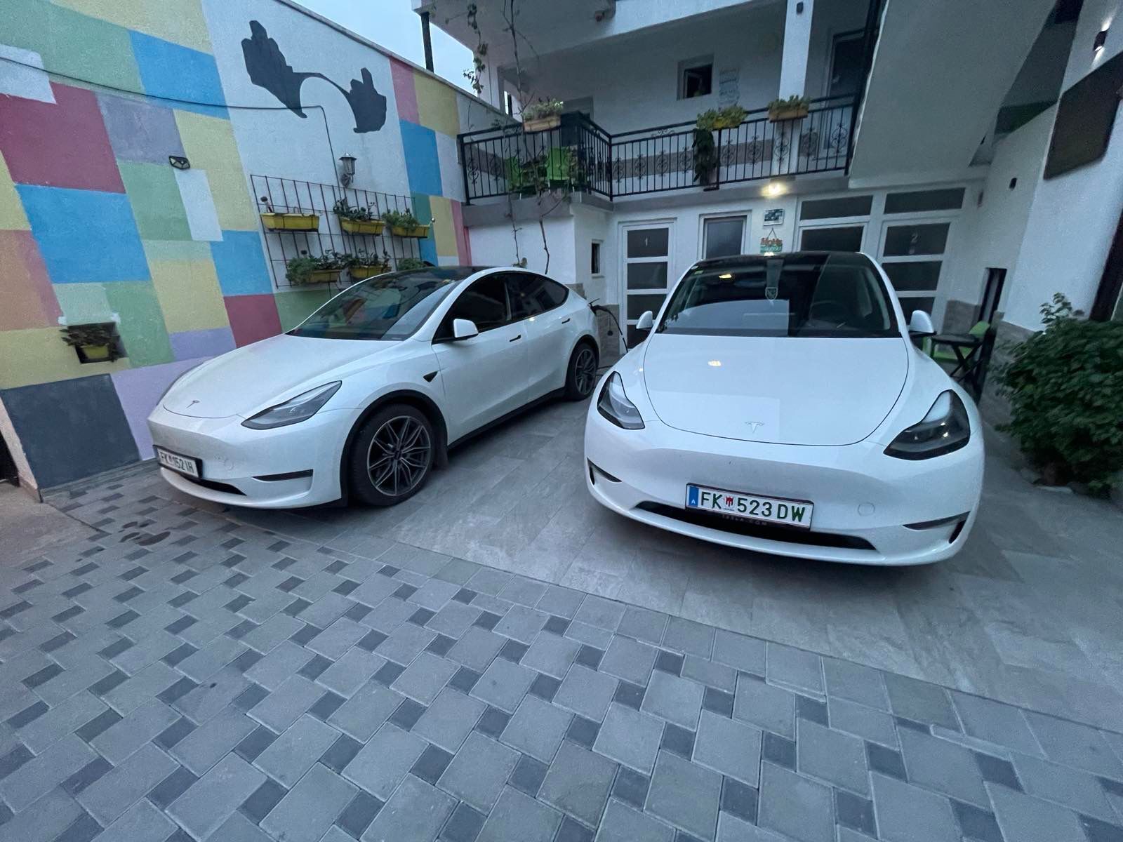 Due auto Tesla bianche parcheggiate in un'area coperta con un muro colorato e un balcone con piante.