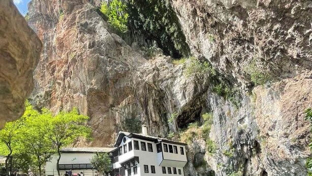 A white building with multiple floors, situated at the edge of a clear blue pool, surrounded by rocky cliffs.