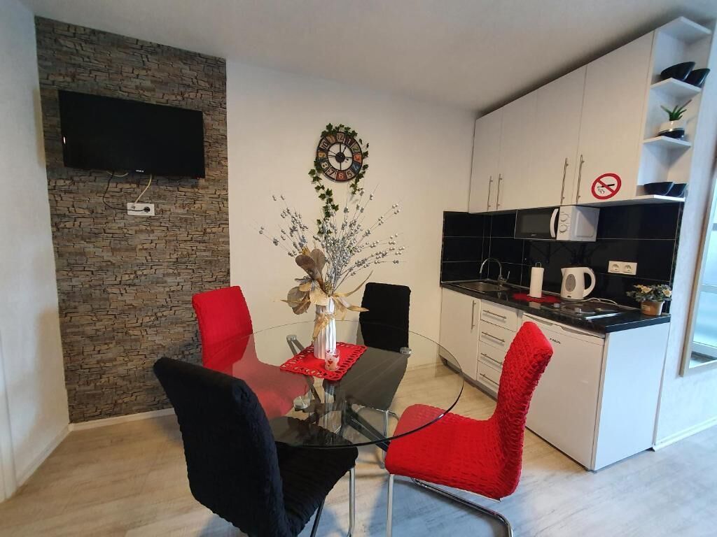 A modern kitchen with white cabinets, a black countertop, and a built-in microwave. There is a red chair and a glass table with a vase of flowers.