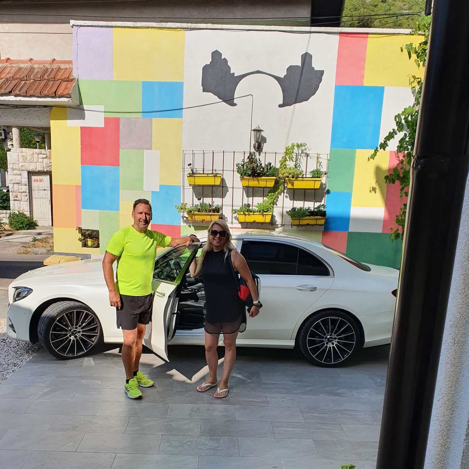 Two people standing in front of a white car with a colorful mural in the background.