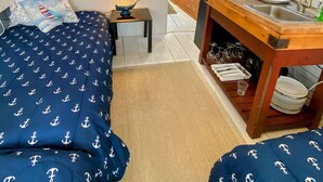 A room with two beds, a small table with a lamp, a kitchen area, and a TV mounted on the wall.