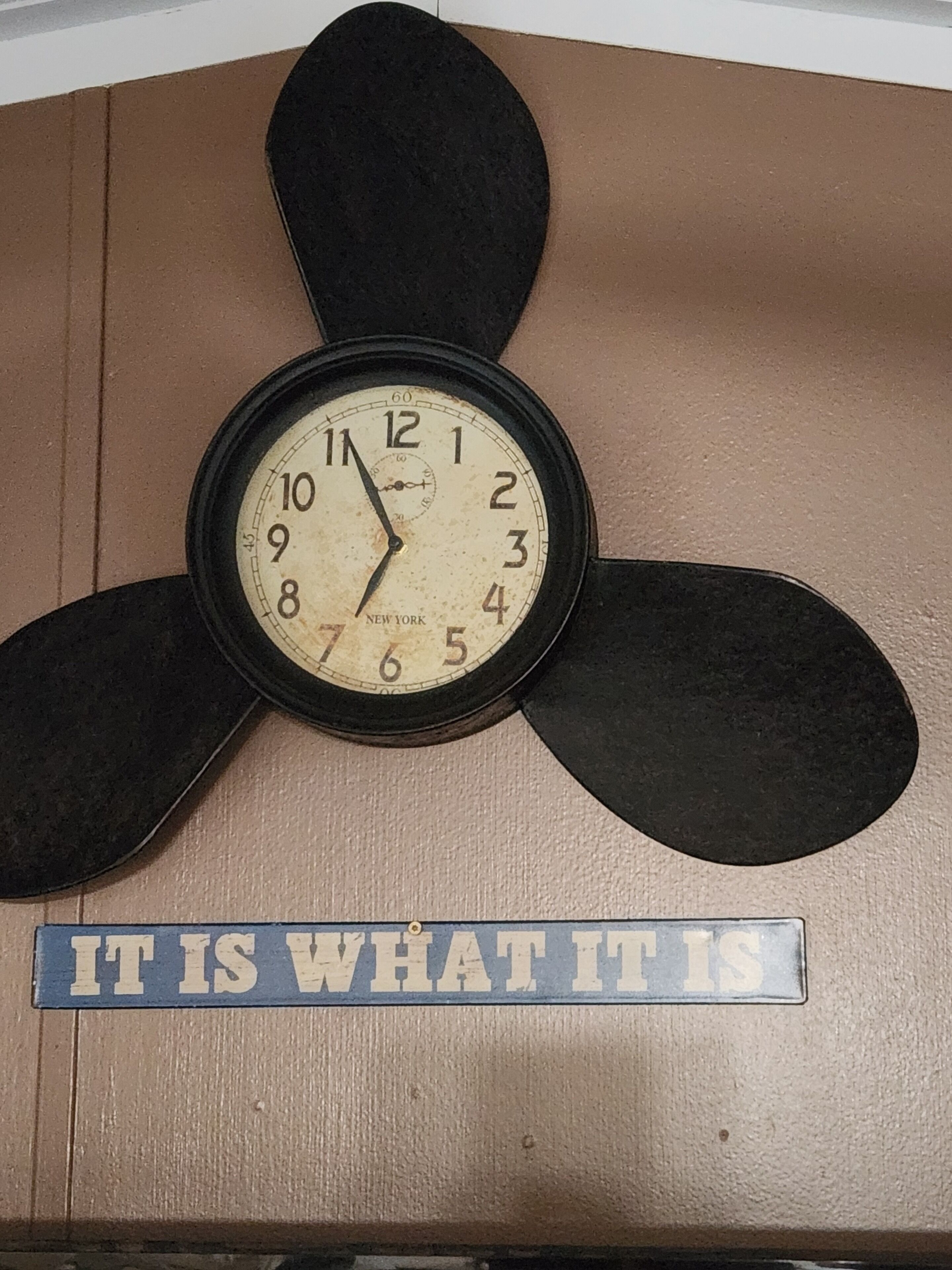 A wall clock with a propeller design and a sign below it that reads "IT IS WHAT IT IS".