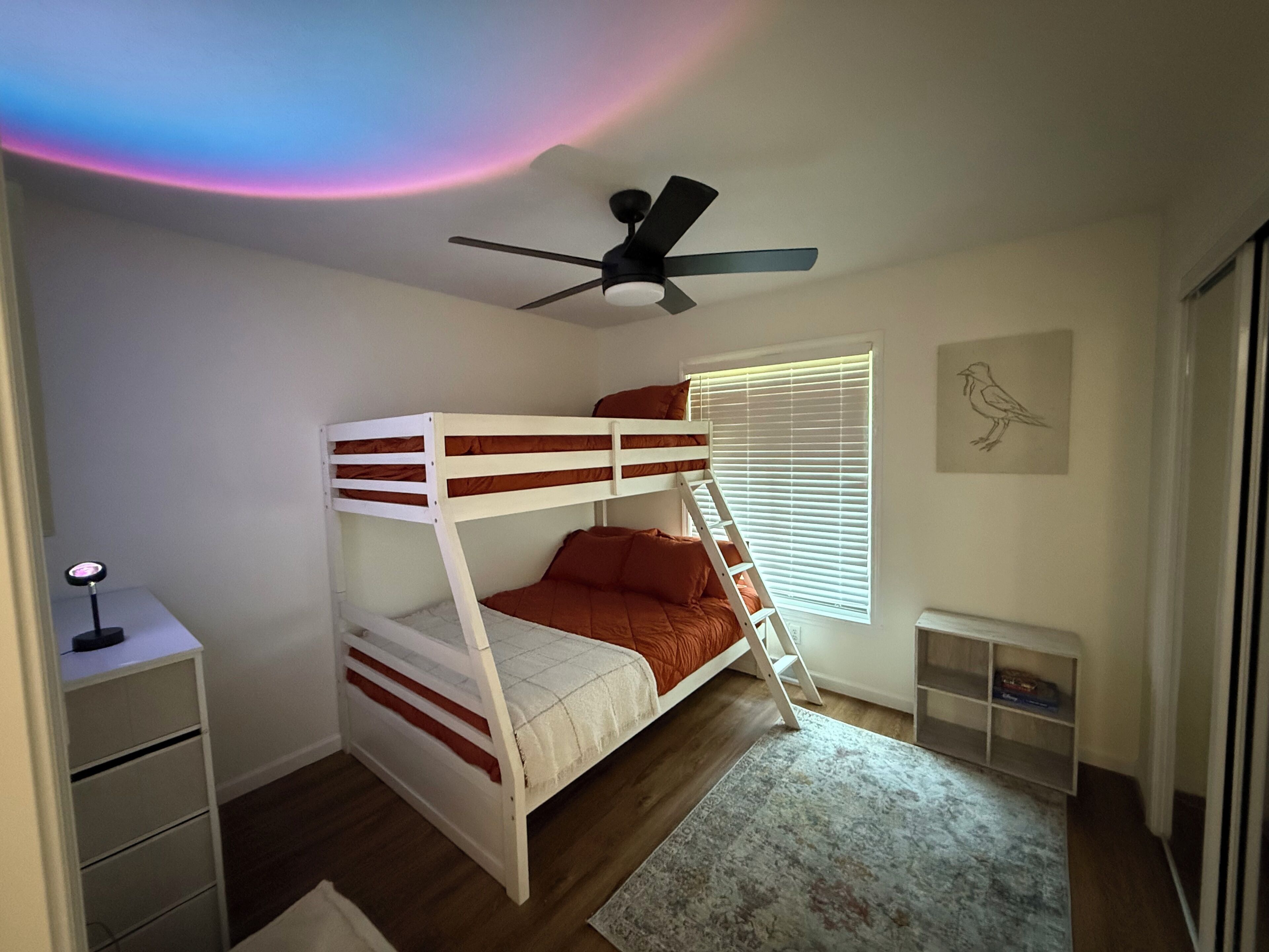 Ein Schlafraum mit Etagenbetten, Deckenventilator, Fenster mit Jalousien, kleinem Schreibtisch und Teppich.