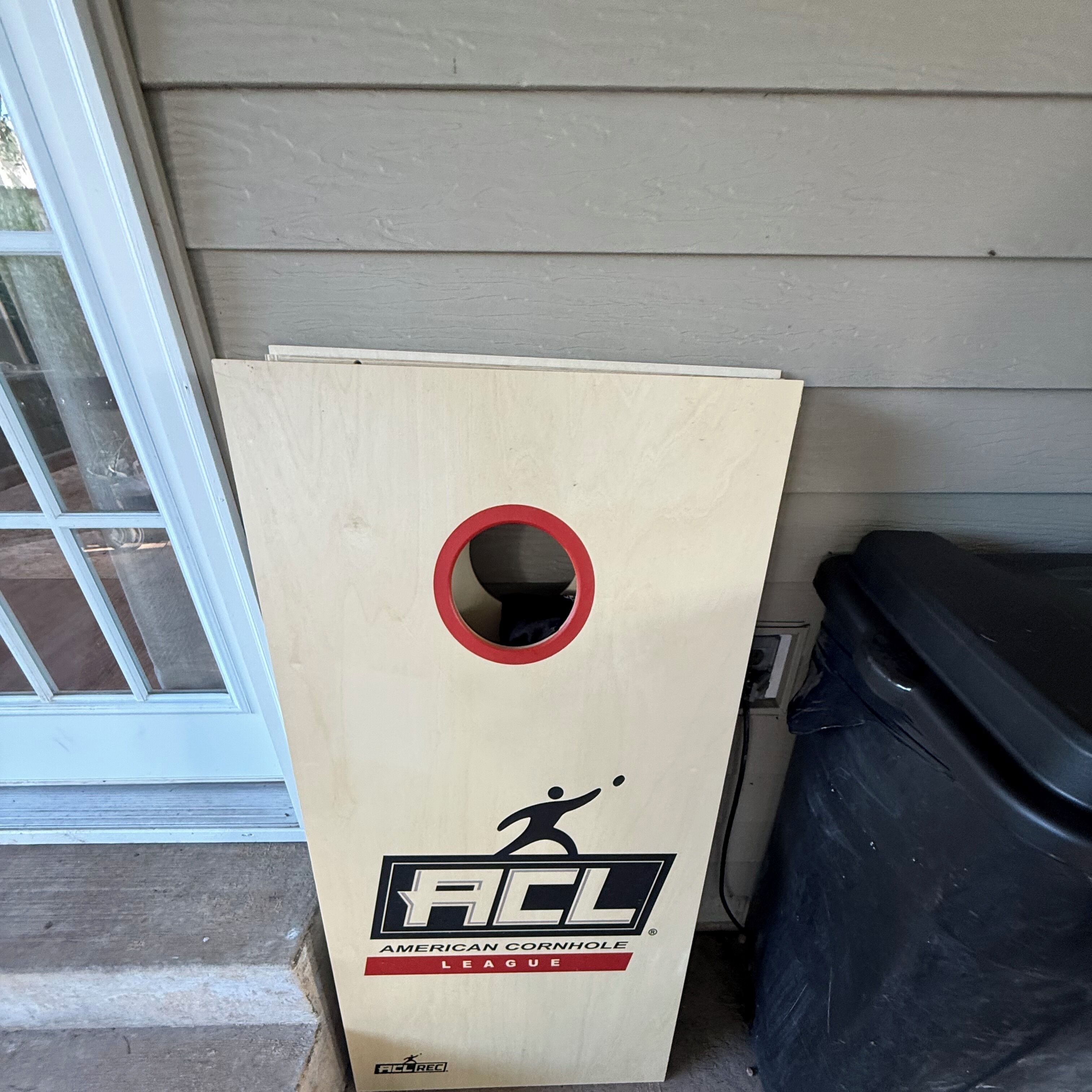 Un sac en tissu de l’American Cornhole League, fait de bois, avec un cercle rouge découpé, placé à l’extérieur d’une maison, à côté d’une poubelle noire.