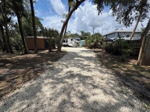 A gravel driveway leading to a mobile home with a porch, surrounded by trees and a wooden fence.