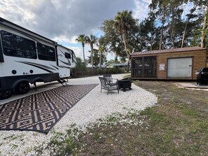 A recreational vehicle parked in a gravel area with a patio table and chairs, surrounded by trees and a wooden structure with a sign.