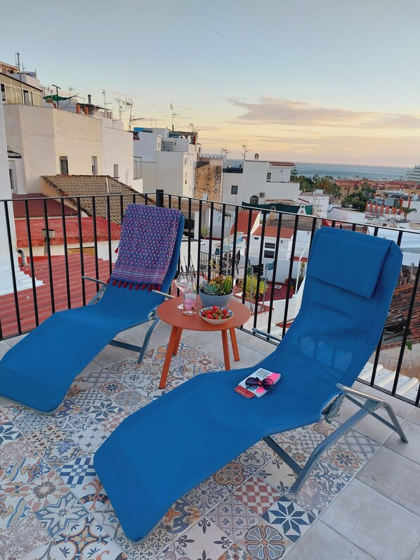 A balcony with two blue lounge chairs, a small table with a bowl of fruit and a bottle, and a view of the cityscape and sea. - Andalusian House with Sea View in the Old Town (Almuñécar)