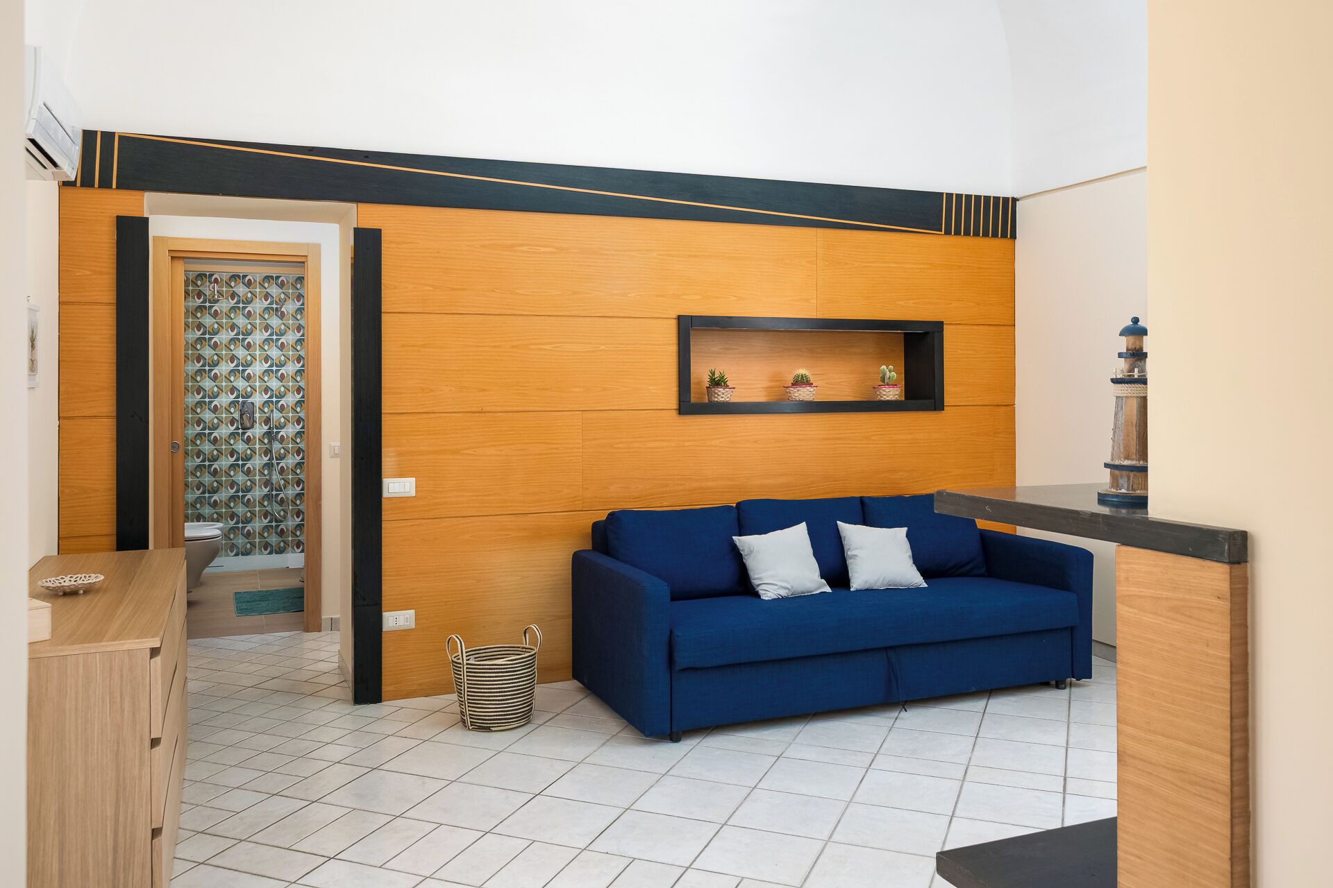 A modern living room with a blue sofa, a small wooden side table, and a wall-mounted shelf with decorative items.