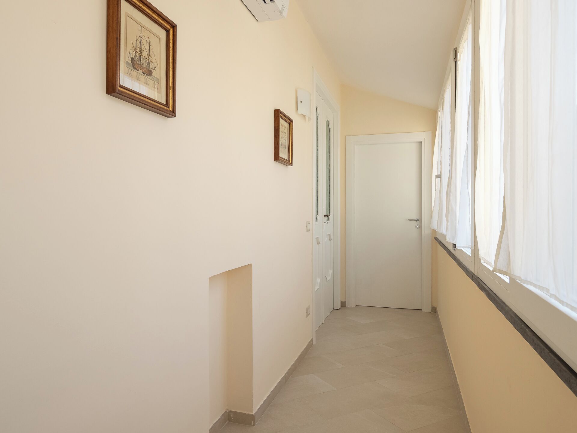 Un couloir étroit aux murs de couleur crème, doté d’un sol carrelé et d’une porte blanche au fond.