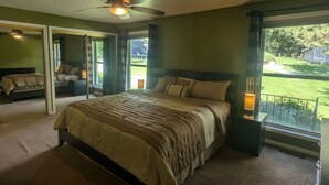 A bedroom with a large bed, a ceiling fan, and a view of a grassy yard through a window.