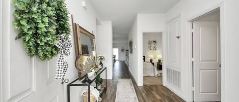 A hallway with a wooden floor, white walls, and a hanging green plant. There is a console table with a globe and a mirror.