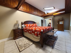 Une chambre avec un plafond en bois, un lit recouvert d’une couvre-lit à motifs rouges et blancs, une table de chevet avec une lampe, une commode et un miroir.