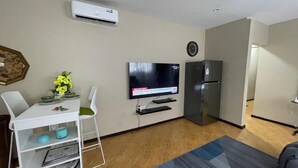 A modern living room with a flat-screen TV, a refrigerator, a dining table with chairs, and a small shelf unit.