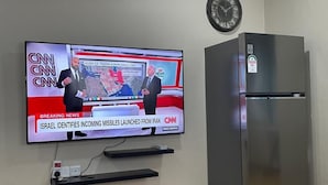 A television displaying a news broadcast, a wall clock, and a stainless steel refrigerator.