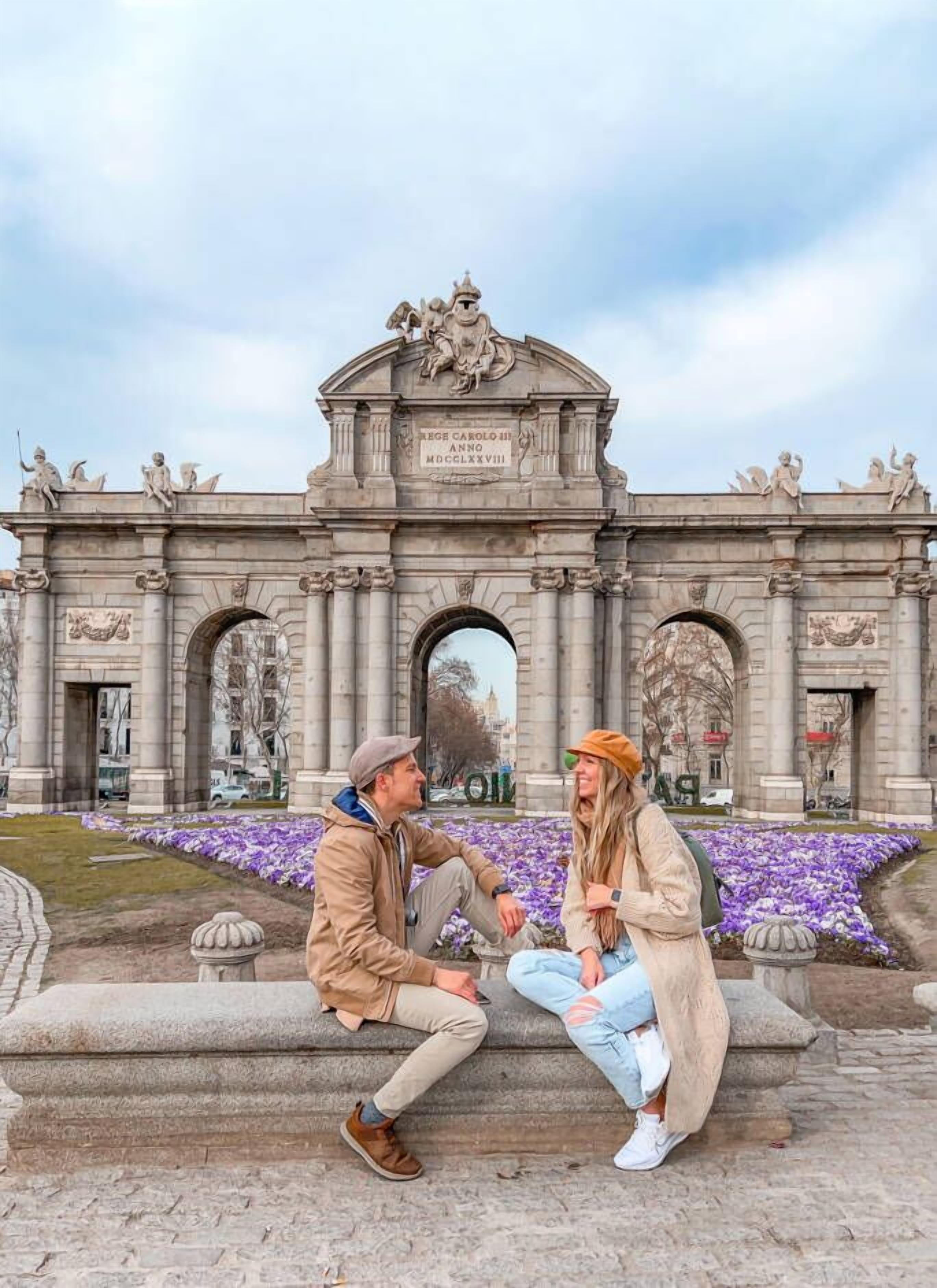 A couple sitting on a stone bench in front of a grand, ornate archway with classical architectural details and a plaque.