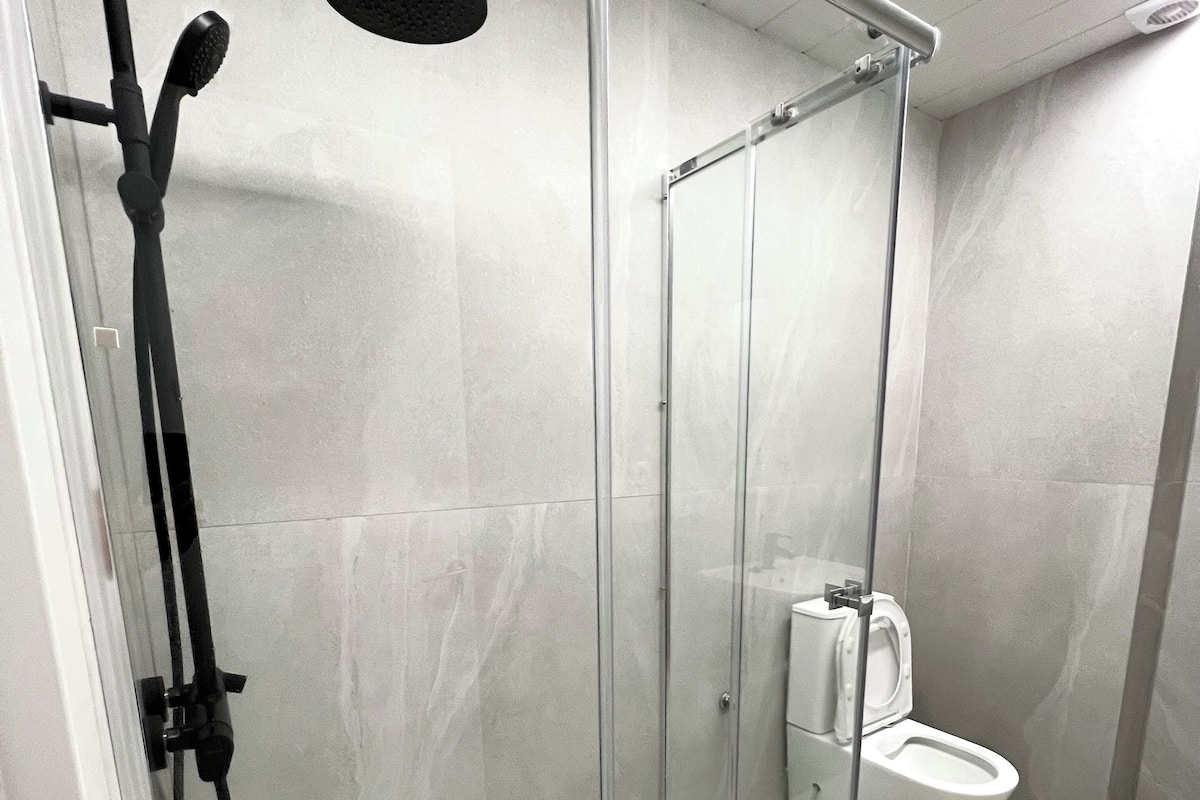 Moderni suihkukaappi, jossa on lasiovi, käsisuihku ja wc, joka näkyy oven läpi.