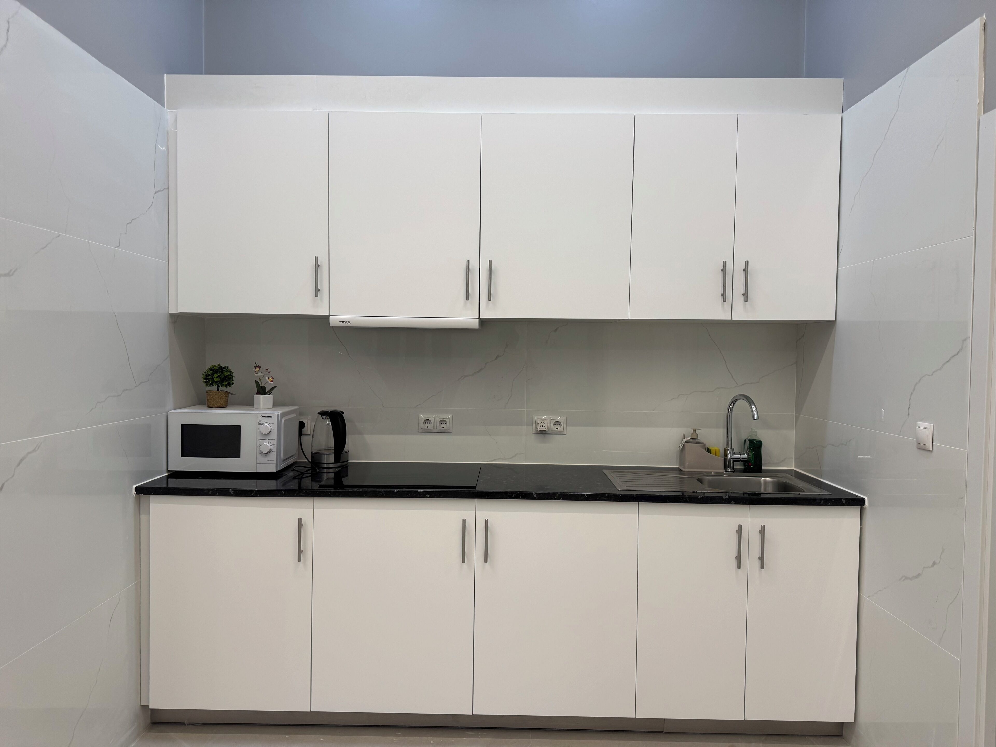 A compact kitchen with white cabinets, a microwave, a kettle, and a sink.