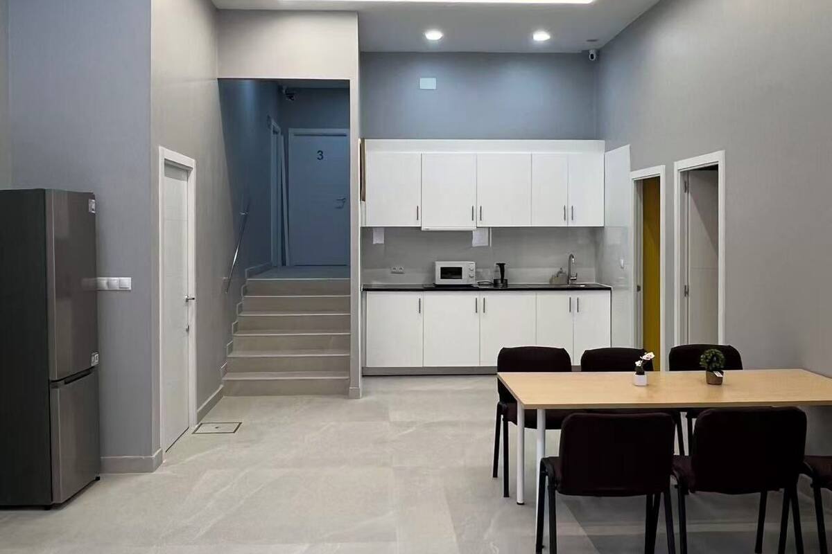 Una cucina moderna con mobili bianchi, un forno a microonde e un frigorifero in acciaio inossidabile. C'è un'area pranzo con tavolo e sedie, e una scala che conduce a un piano superiore.