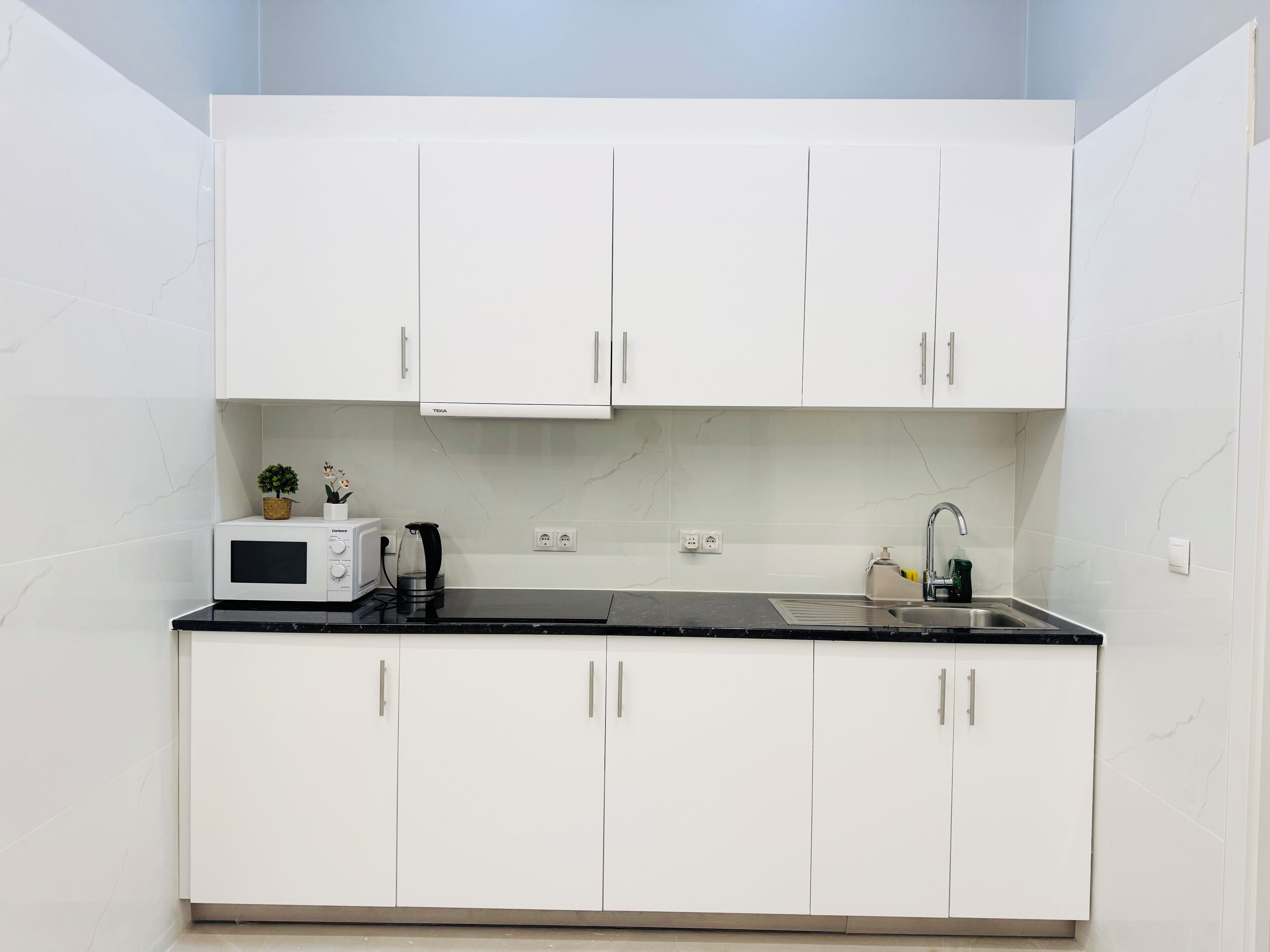 A compact kitchen with white cabinets, a microwave, a kettle, and a sink.
