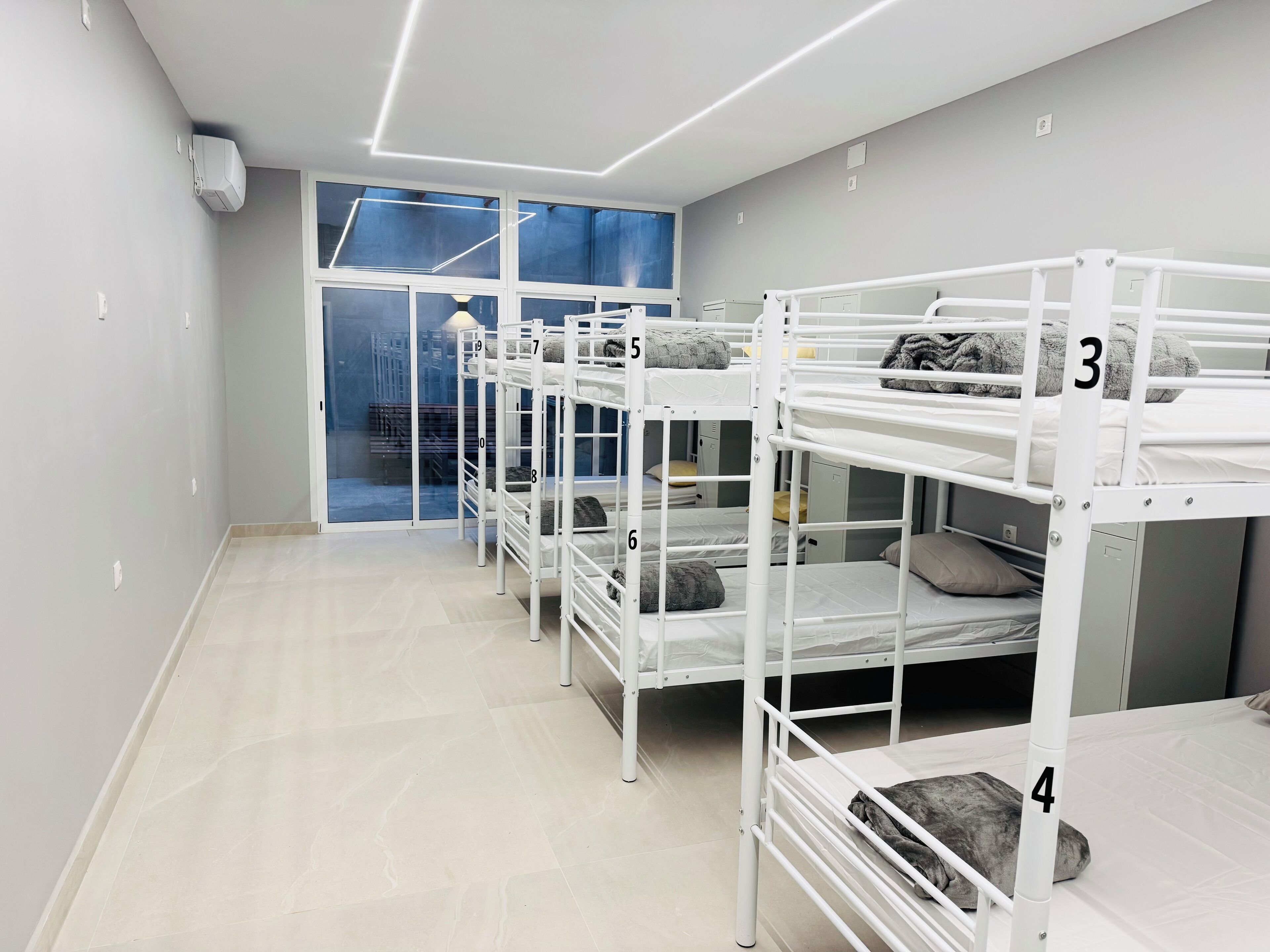 A dormitory room with bunk beds numbered 3 to 9, a window with blue curtains, and a white ceiling with linear lighting.