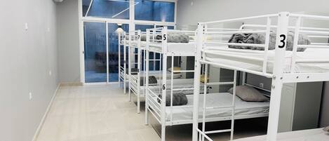 A dormitory room with bunk beds numbered 3 to 9, a window with blue curtains, and a white ceiling with linear lighting.