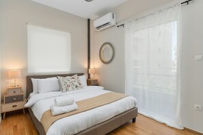 Un dormitorio con cama, mesitas de noche, equipo de aire acondicionado, ventana con cortinas traslúcidas y espejo montado en la pared.