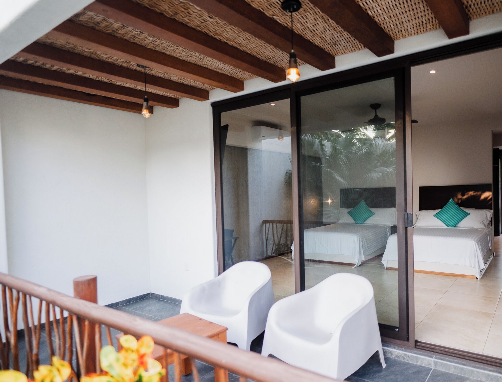 A balcony with two white chairs, a wooden table, and a glass door leading to a bedroom with a bed and two pillows.