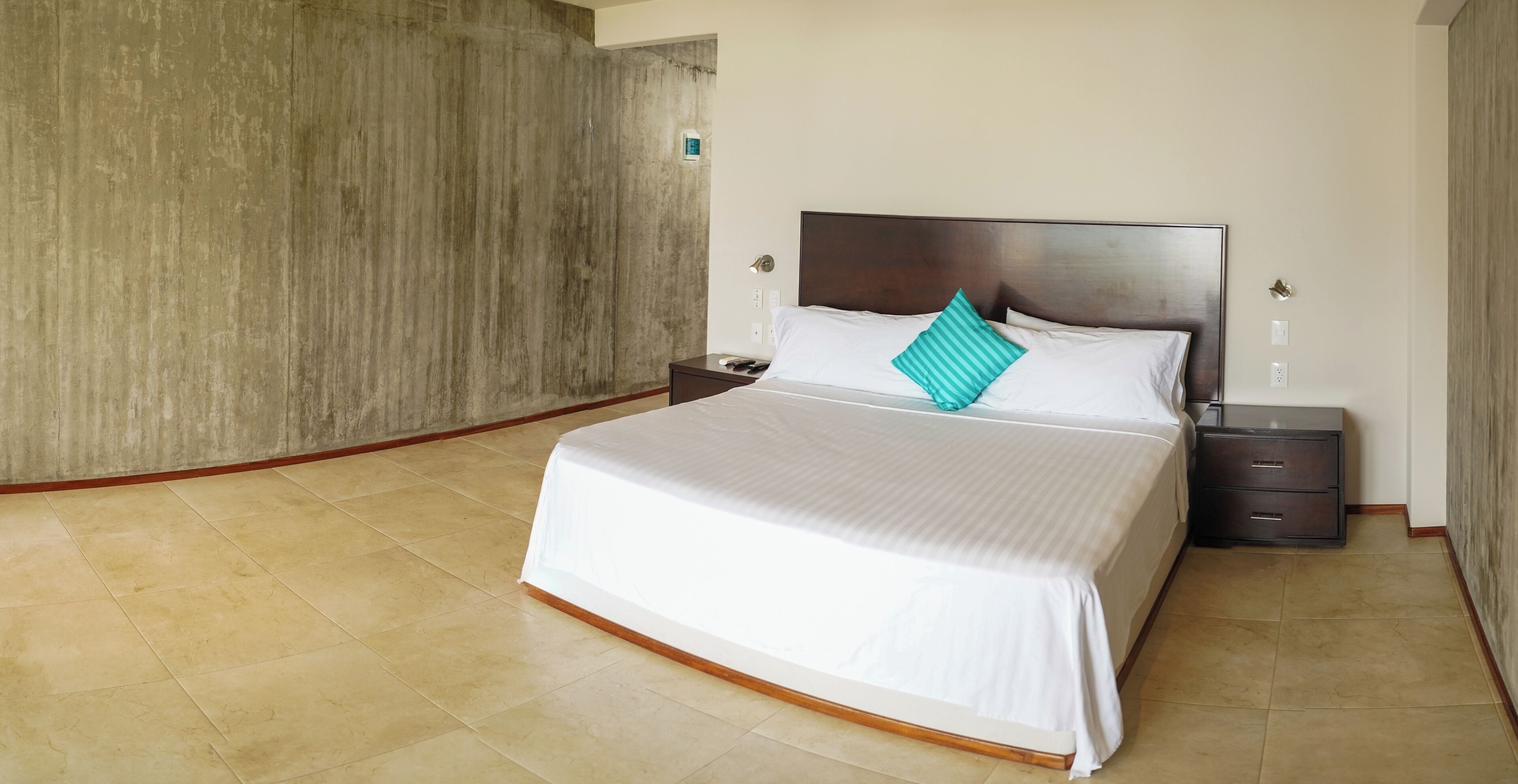 A hotel room with a large bed, a nightstand, a drawer, and a concrete wall.