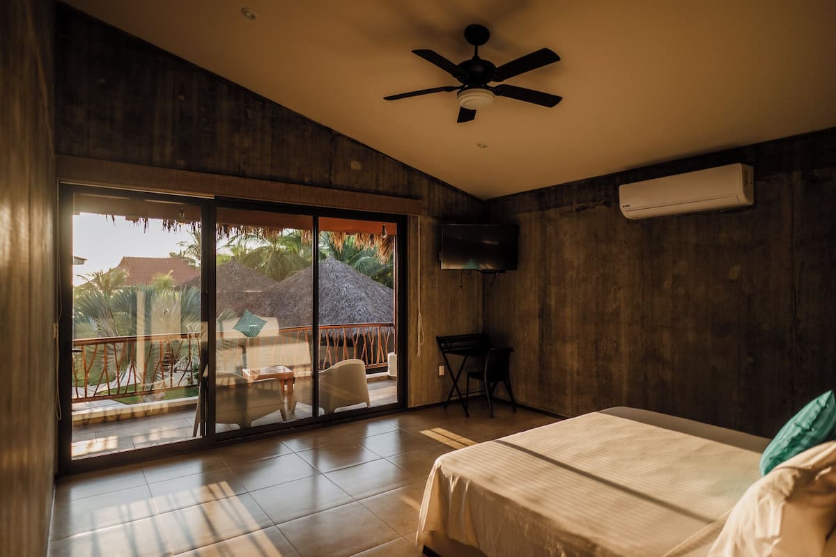 A bedroom with a bed, a ceiling fan, a TV, and a balcony with a view of palm trees and a thatched roof structure.