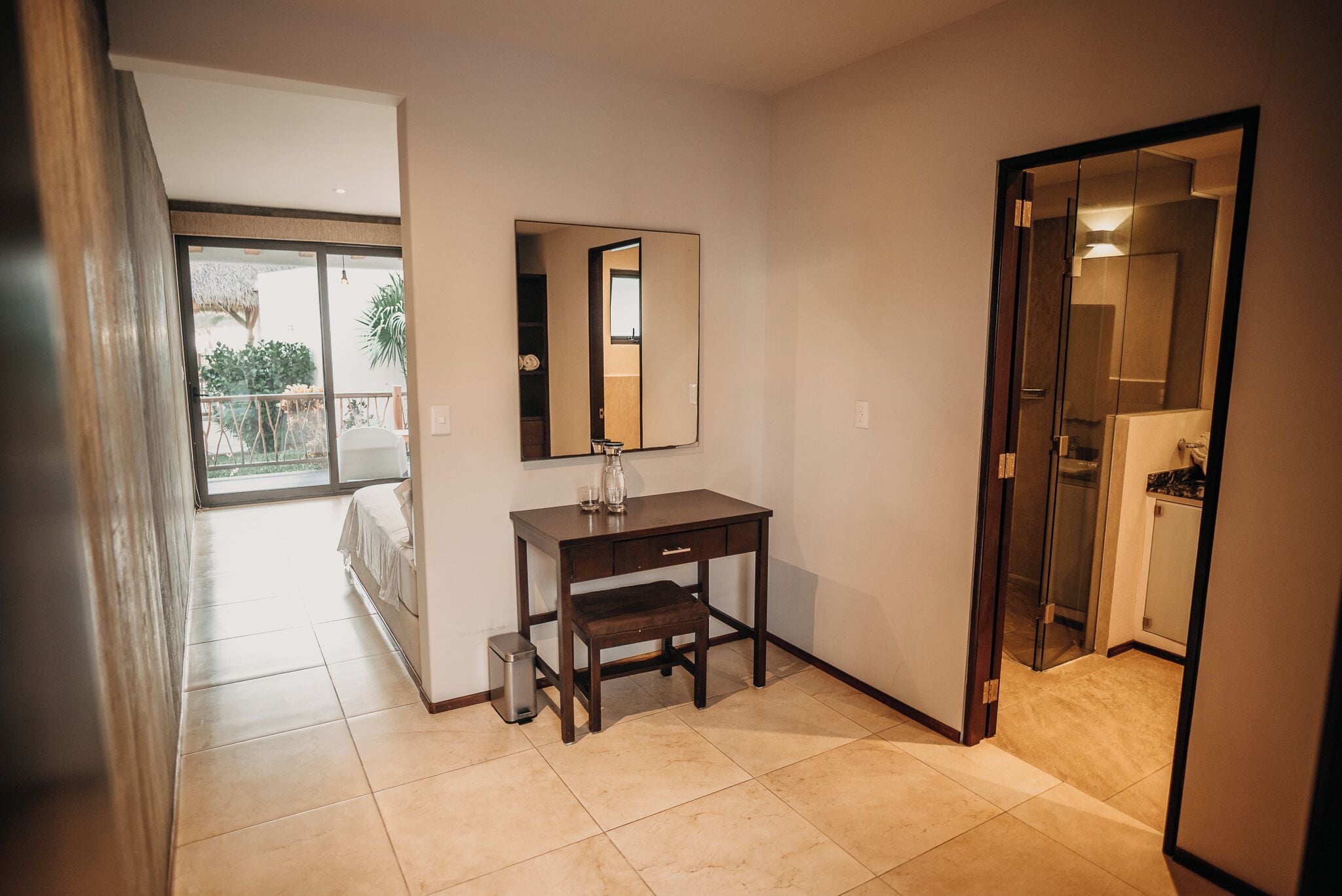 Habitación de hotel con un amplio ventanal corredero que da a un balcón, un escritorio de madera oscura con un espejo y un pequeño cubo de basura.