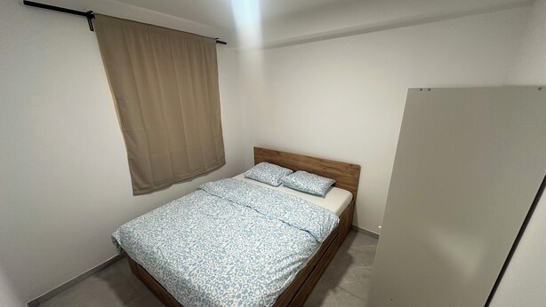Una piccola camera con un letto singolo, una testiera in legno, una tenda beige e una porta bianca.