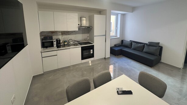Una cucina compatta con mobili bianchi, un forno a microonde integrato e un piano cottura a gas. Un tavolo da pranzo con sedie si trova al centro. In zona soggiorno, un divano grigio e una finestra con vista.