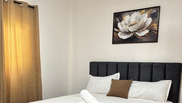 Una cama bien tendida con sábanas blancas y almohada marrón, cabecera oscura y un cuadro con flores en la pared.
